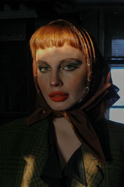 Young trendy woman with red lips and piercings in checkered jacket looking at camera in sun rays indoors