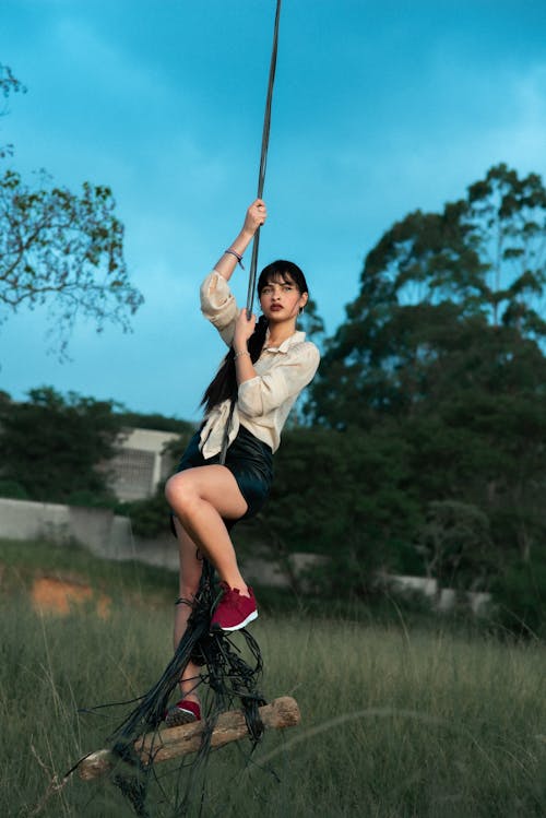 Full body of stylish female in sneakers swinging on rope with wooden plank in suburb area against tree and structure
