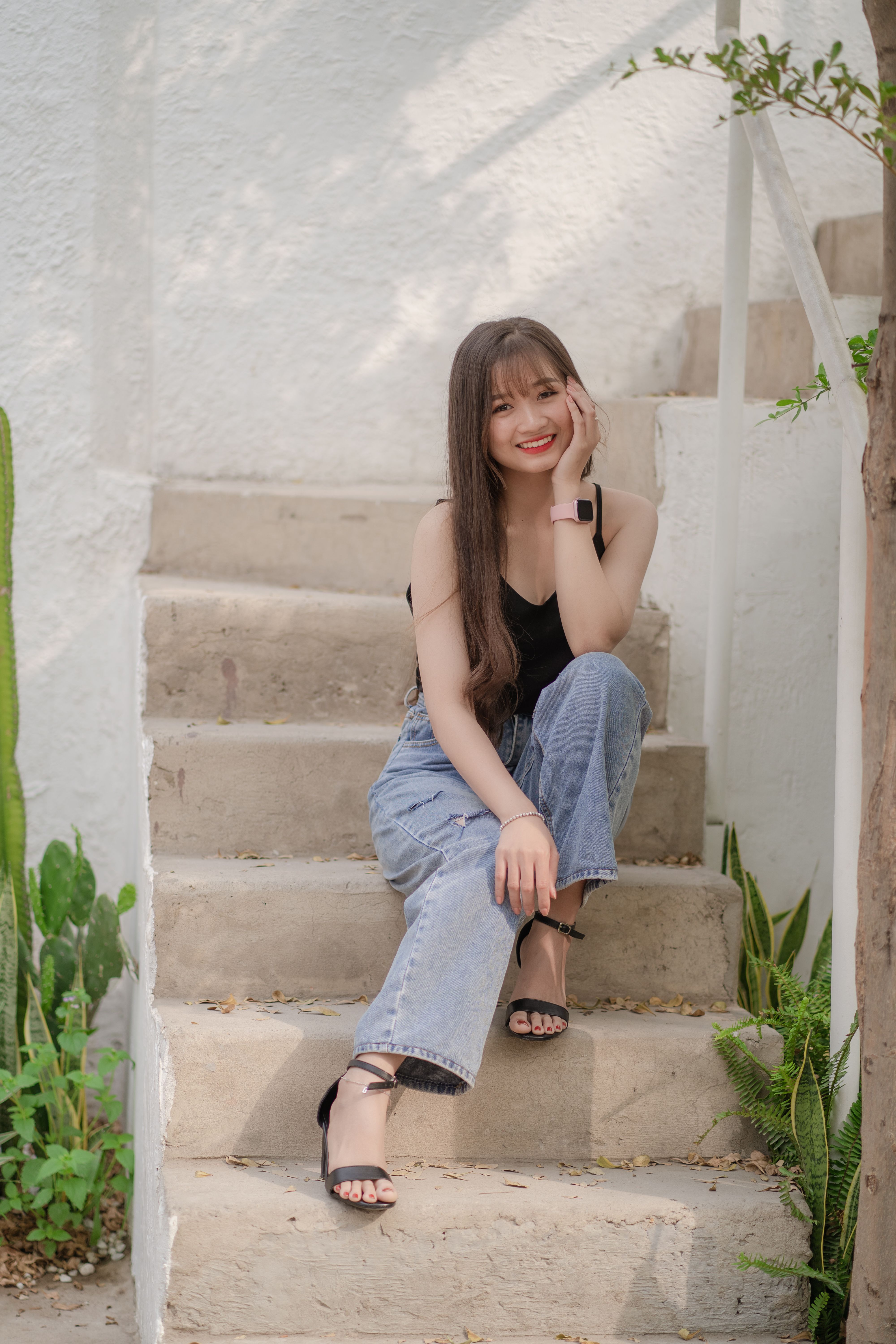 A young woman in casual denim and black top sitting on outdoor concrete stairs, smiling.