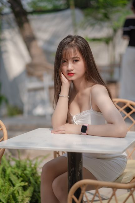 Asian woman sitting at an outdoor cafe in a white dress, looking elegant and relaxed.