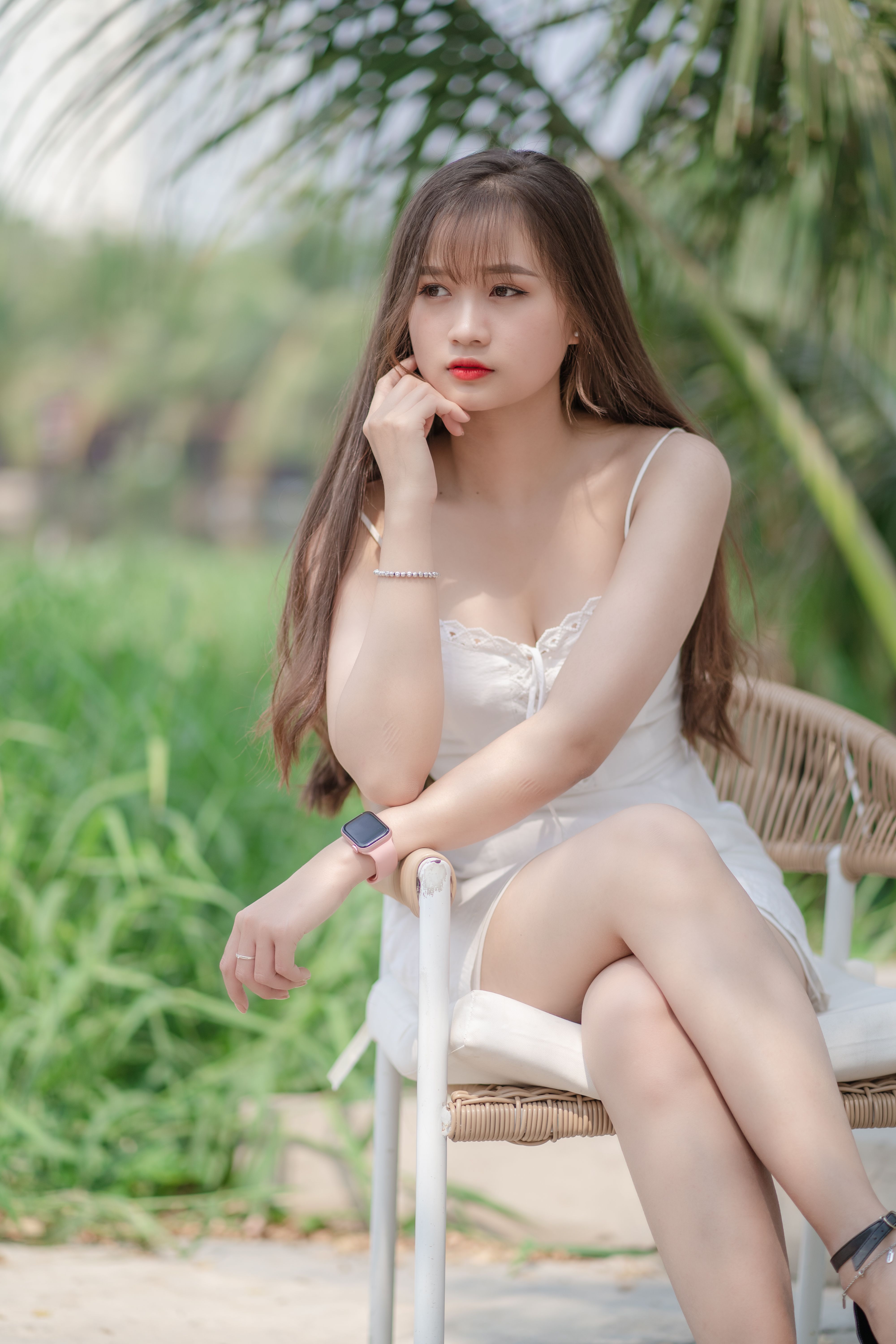 Stylish young woman posing in a white dress on an outdoor chair with a serene background.