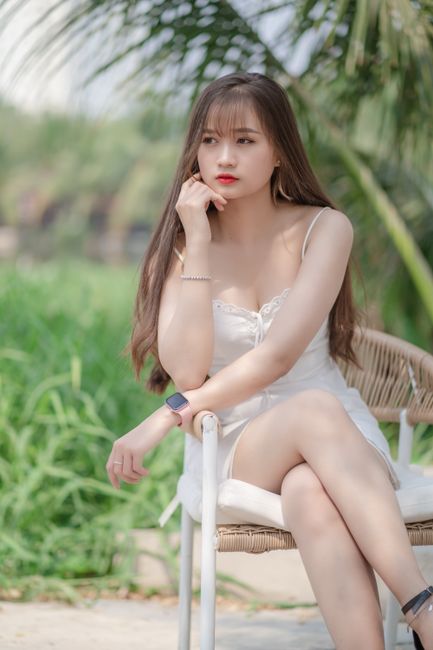 Stylish young woman posing in a white dress on an outdoor chair with a serene background.