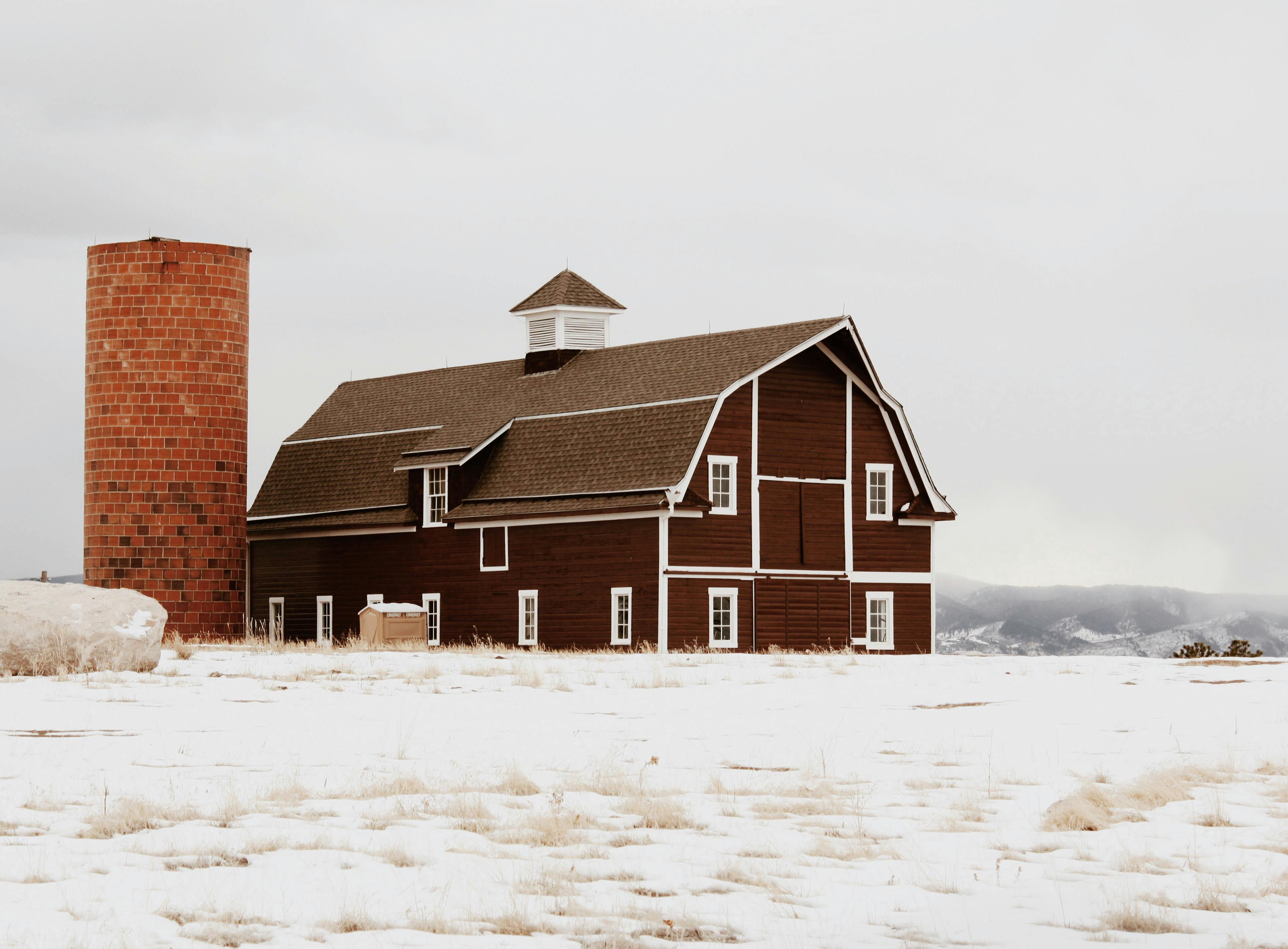 brown wooden barn and silo with brick wall on snow covered ground