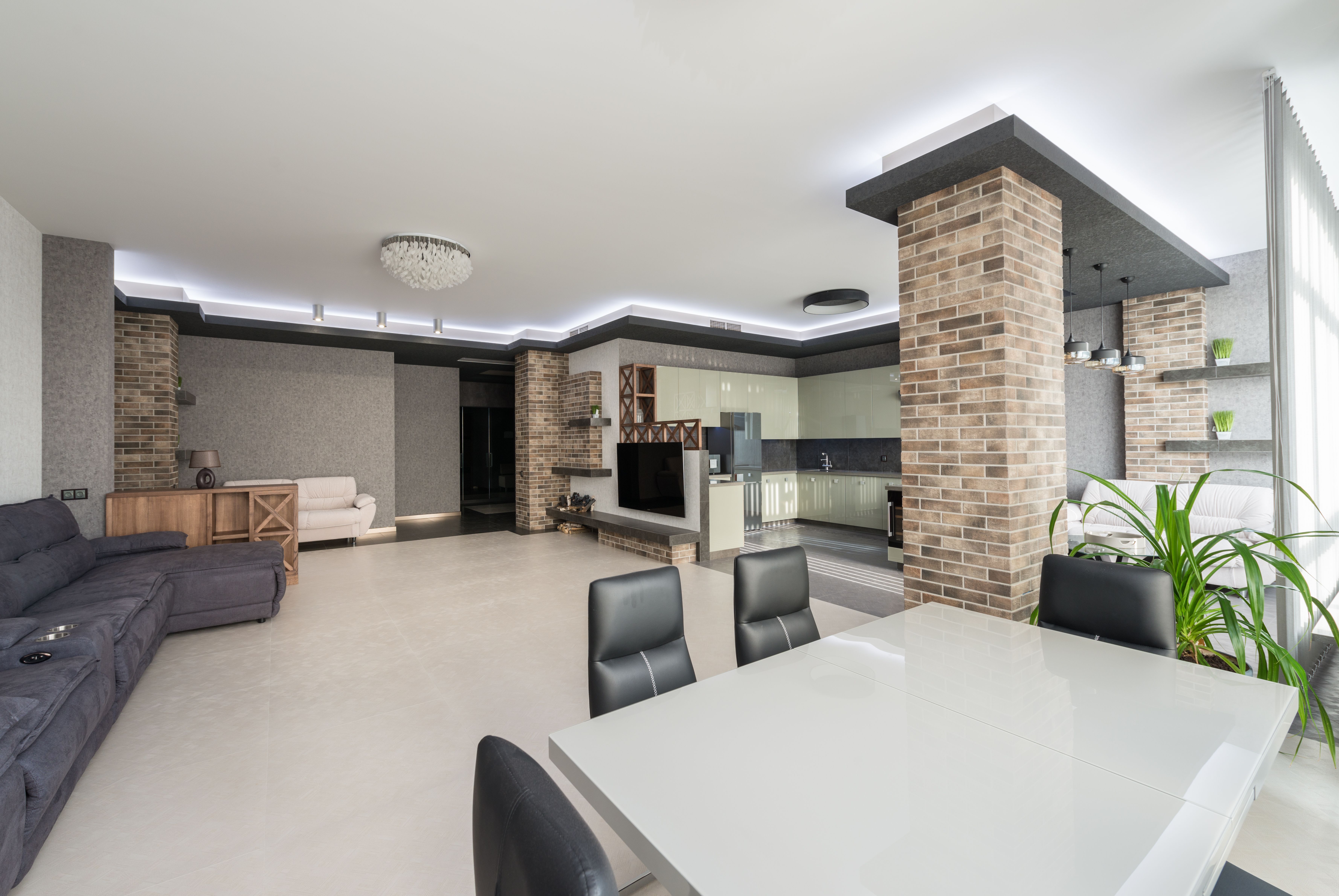 White table with chairs placed in spacious living room with comfortable couch and TV in modern apartment with kitchen and brick column