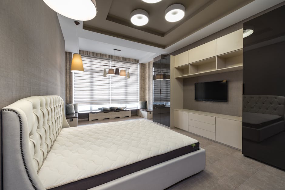 Firm vs Medium Mattress: Back Pain Support Showdown Bed with white mattress placed against wall with cupboards and modern TV in spacious bedroom with window and glowing lamps