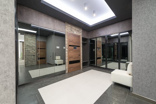 White pouf placed at gray wall in spacious hallway of stylish light apartment with wooden door and mirrors with reflection