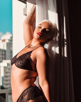 Confident woman in lace lingerie posing by the window with cityscape view.