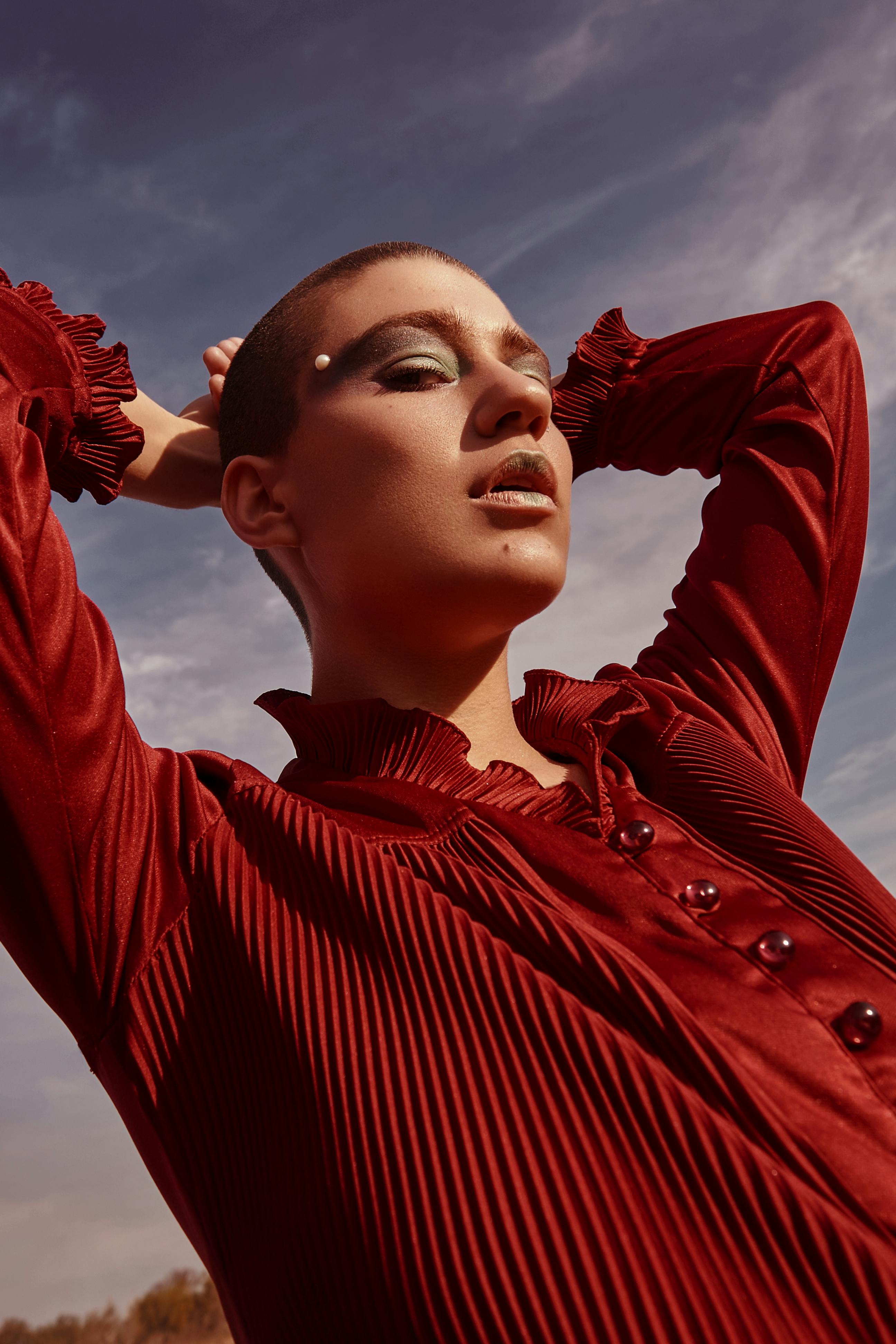 A striking fashion portrait of a model in red with bold makeup, shot from a low angle outdoors.