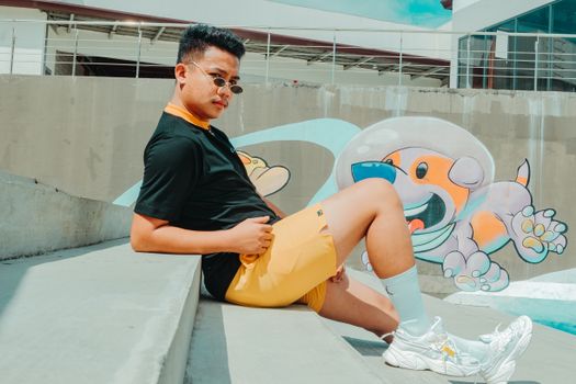 Full body side view of confident ethnic male in summer outfit sitting on staircase near graffiti and looking at camera