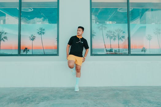 Full body of confident ethnic male in activewear leaning on wall with bog windows decorated with pictures of tropics and looking away