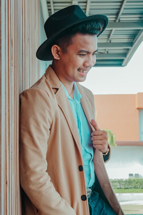 Positive well dressed Asian male in stylish outfit and hat standing near metal wall and smiling happily