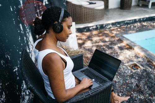 Free Woman sitting by the pool with headphones, working on a laptop in a relaxed outdoor setting. Stock Photo