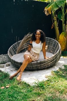 Smiling woman relaxing in a round chair outdoors with her phone.