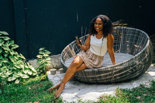 Black woman sitting outdoors on a round chair, listening to music with headphones and holding a smartphone.