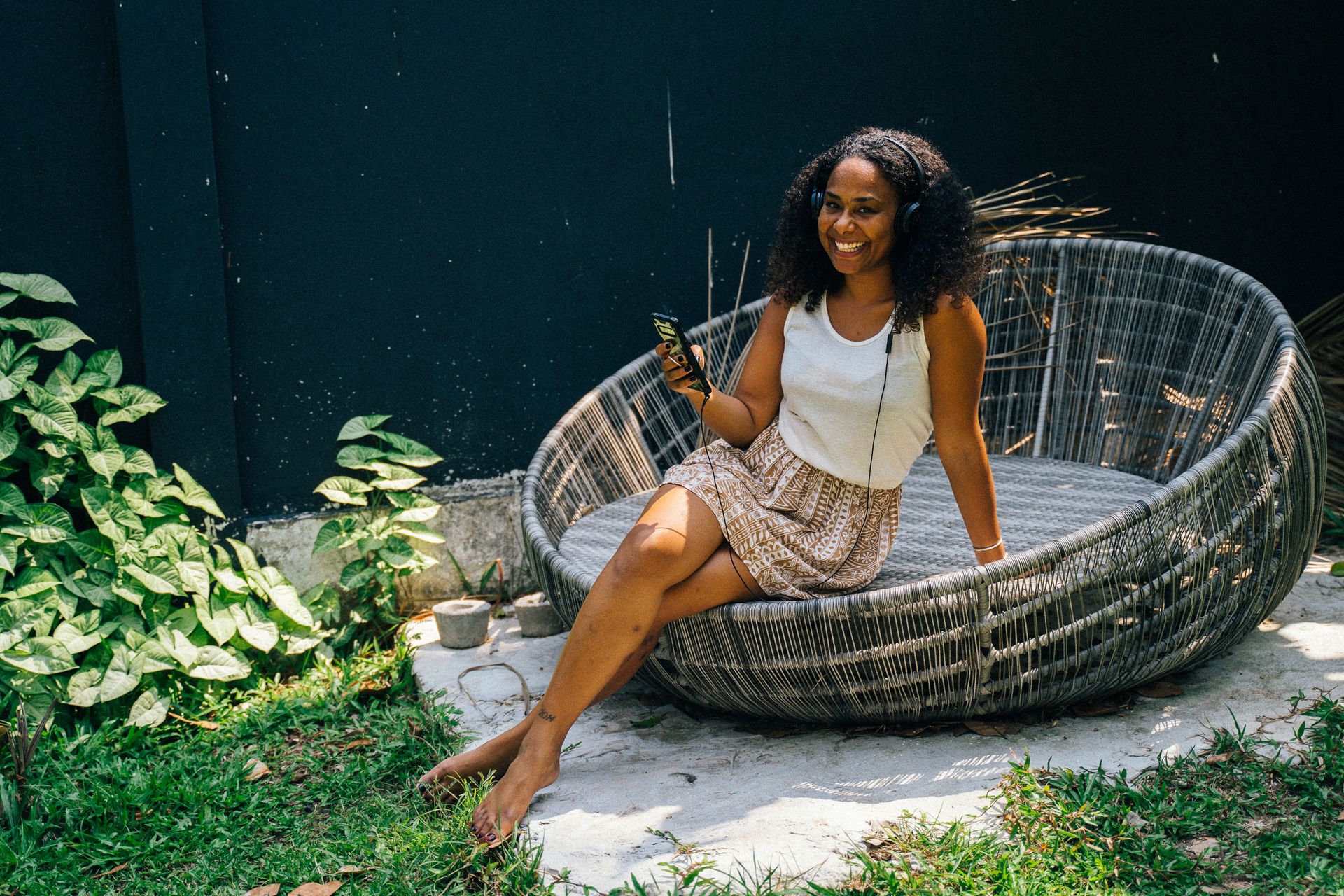 Black woman sitting outdoors on a round chair, listening to music with headphones and holding a smartphone.