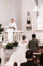 Groom with bride and guests listening to speech of priest standing behind podium in church during wedding ceremony