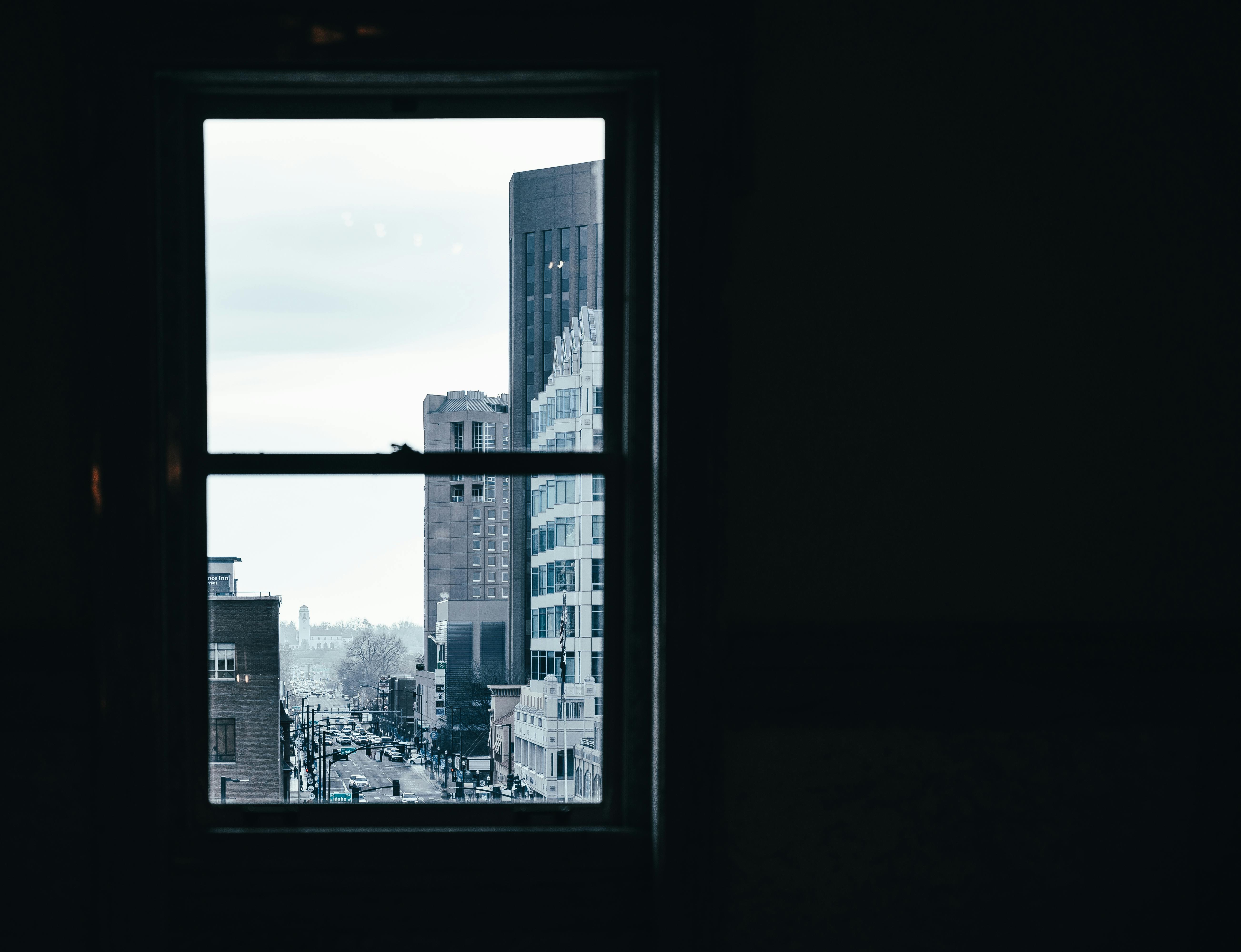 Window in room with dim light overlooking contemporary city with modern multistory buildings and road on street against cloudless sky