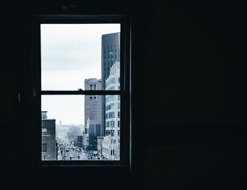 Window in room with dim light overlooking contemporary city with modern multistory buildings and road on street against cloudless sky