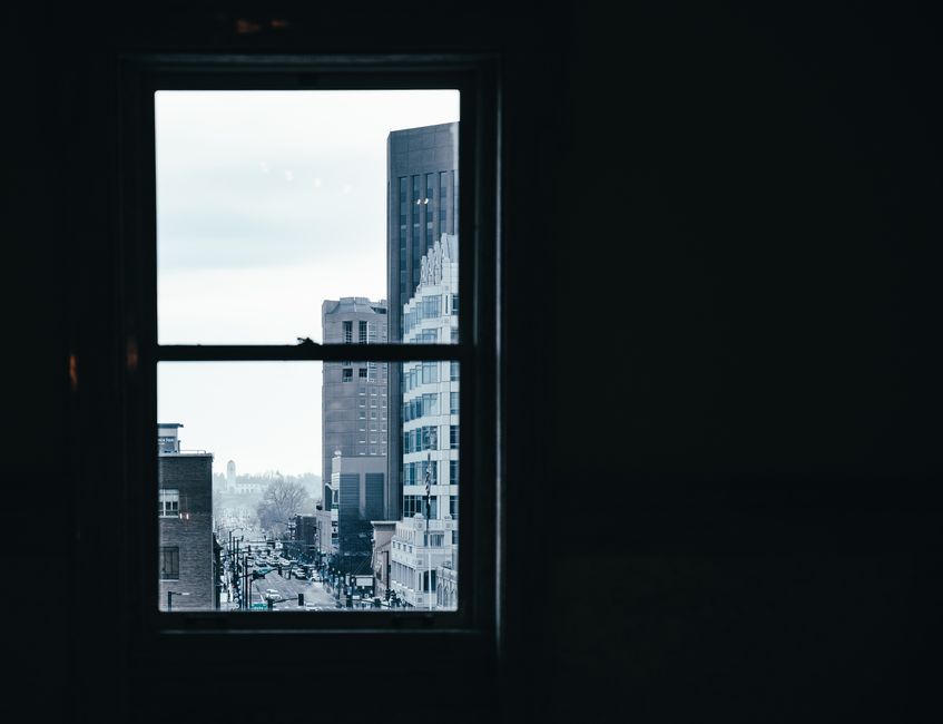 Window in room with dim light overlooking contemporary city with modern multistory buildings and road on street against cloudless sky
