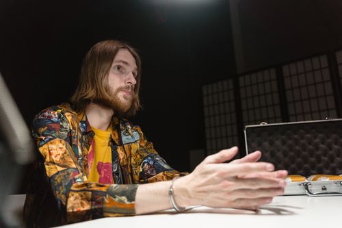 Free A thoughtful man in a patterned shirt sits indoors under dramatic lighting, gesturing with hands. Stock Photo