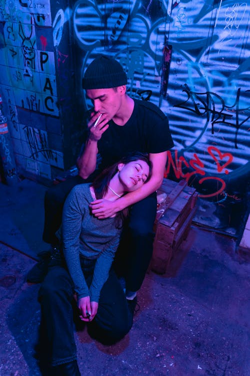 A Woman Sleeping on the Floor while Leaning on a Man Smoking Cigarette