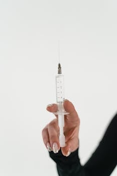 Closeup image of a hand holding a syringe with a visible needle on a light background.