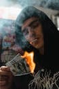 Man in Black Hoodie Smoking a Cigarette and Holding a Burning Paper Bills