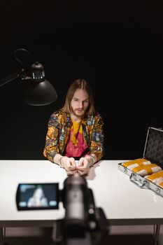Man in patterned jacket seated at table with camera filming, lit by desk lamp.
