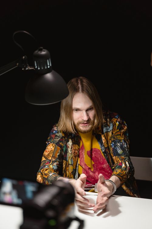 Free A man speaking under a desk lamp, with a colorful patterned shirt in a dim room. Stock Photo