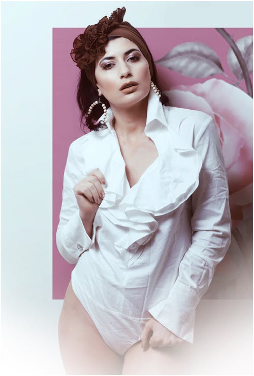 Young attractive female model in white outfit with headband and earrings standing near wall with picture while looking at camera