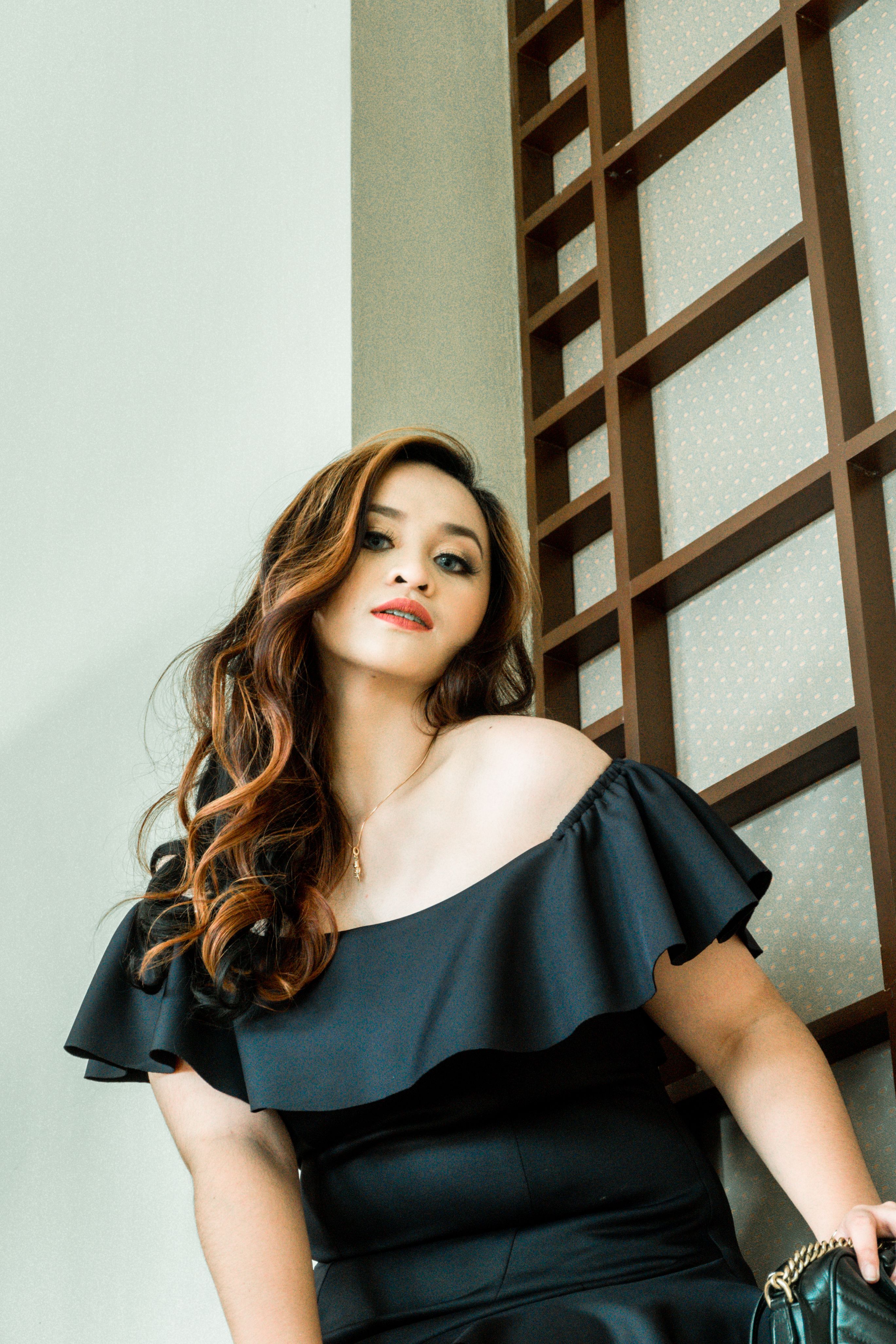Stylish woman with long hair in an off-shoulder black dress posing against a modern interior wall.