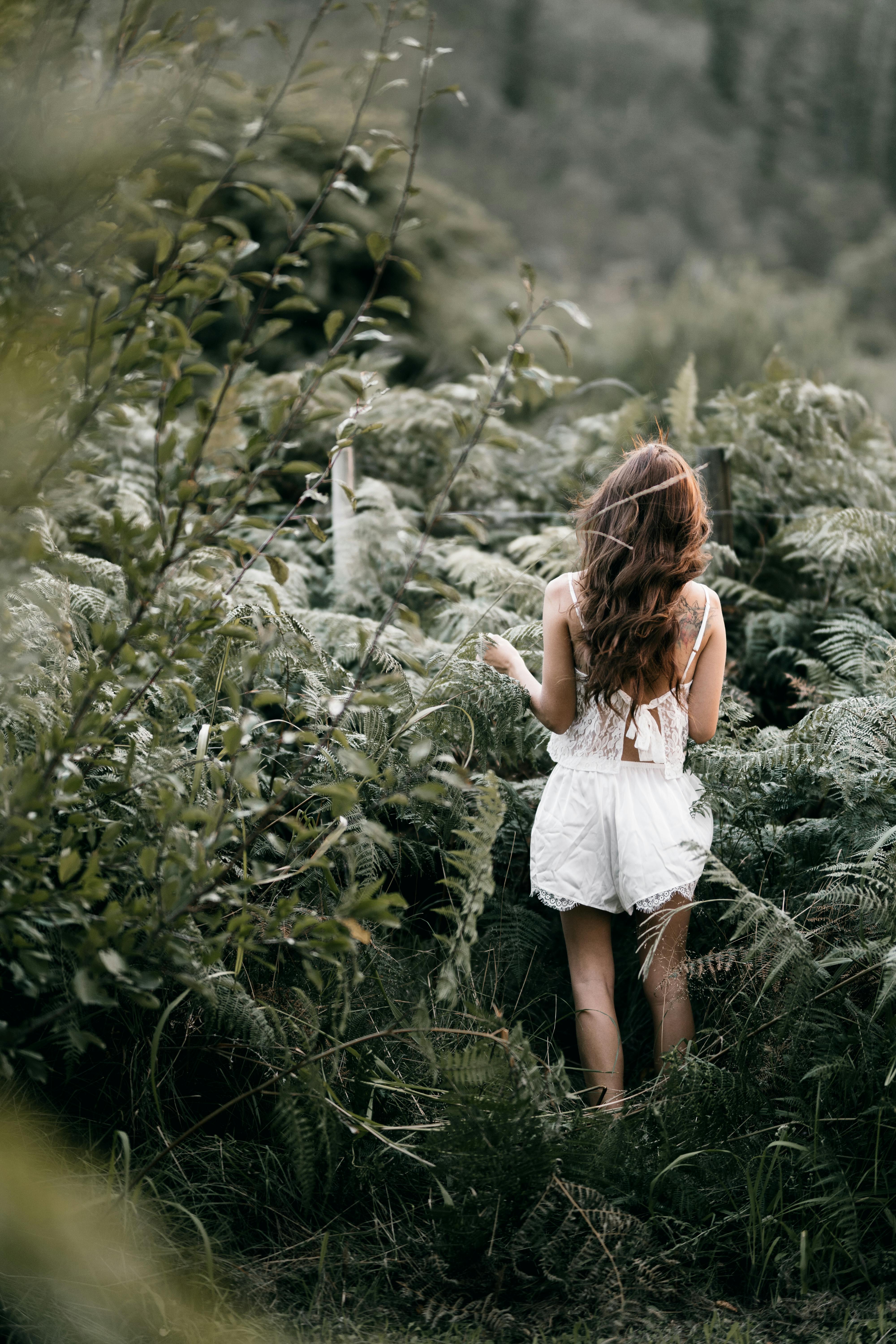 A woman in white clothing stands amidst lush greenery, embracing nature's tranquility.