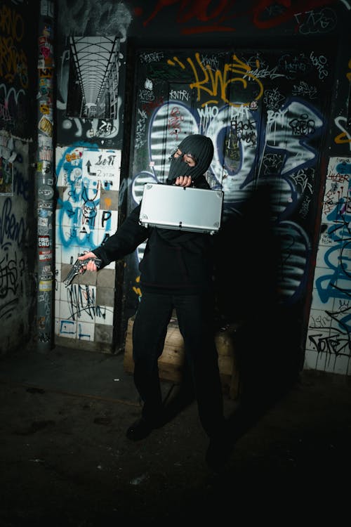 Free Person in Black Jacket with Mask Holding a Silver Briefcase and Gun Stock Photo