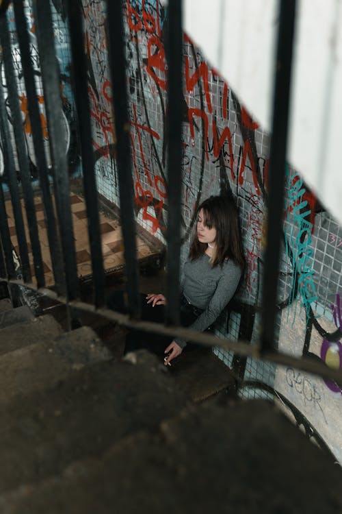Woman in Gray Long Sleeve Shirt Sitting on Concrete Stairs and Smoking