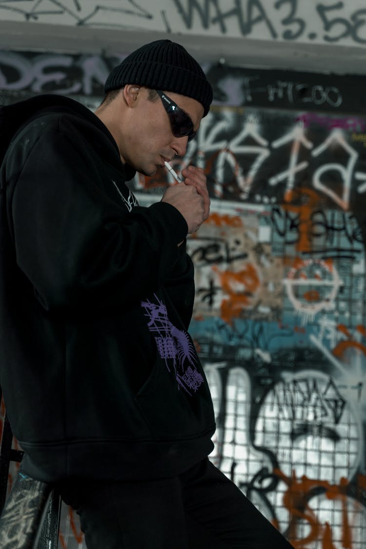 A Man Wearing A Beanie And Shades Lighting A Cigarette