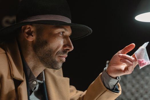 A man in a coat and hat holds a bag of red pills under a spotlight.