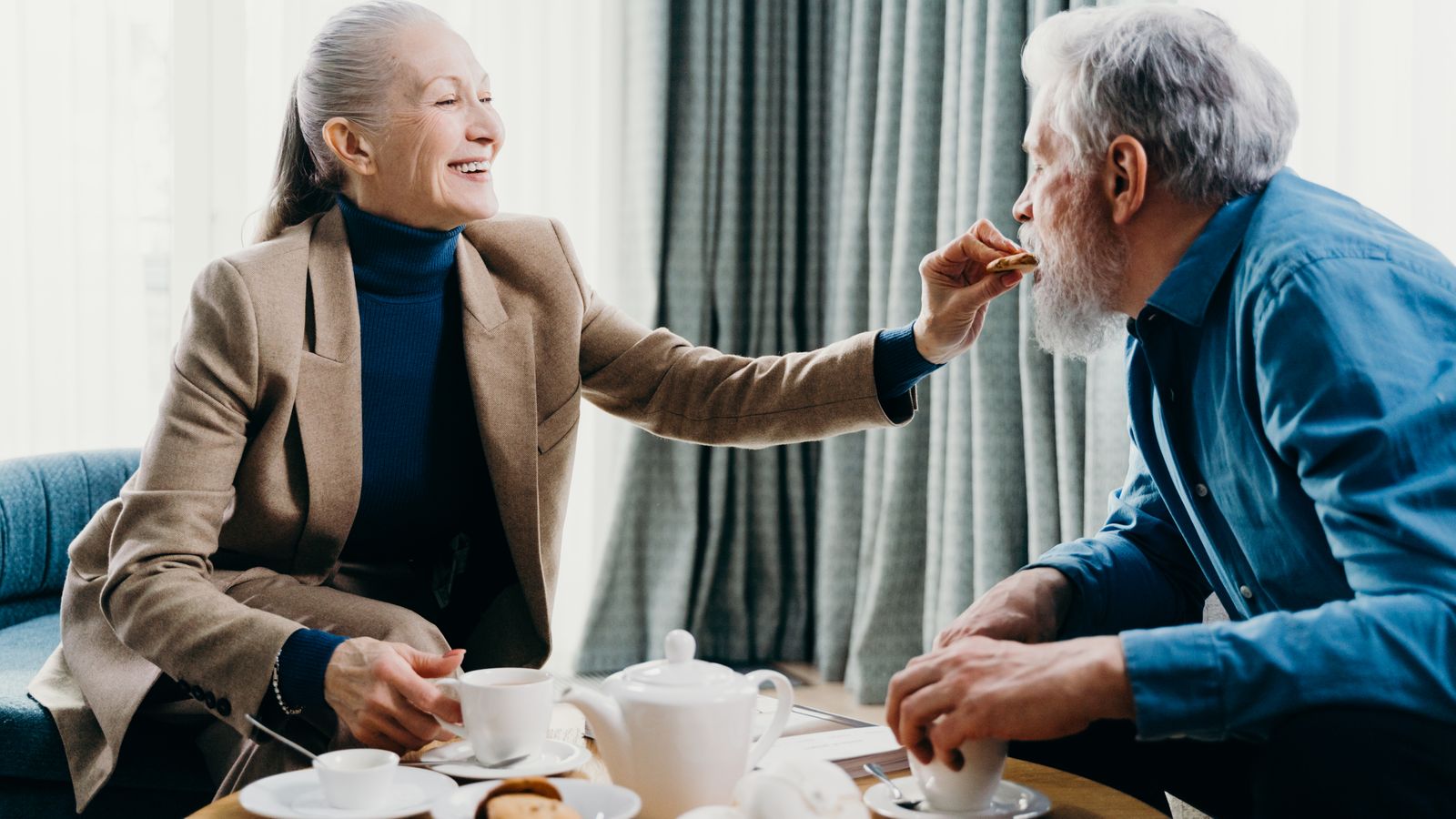 Two older adults sharing tea and a biscuit at a table by a window