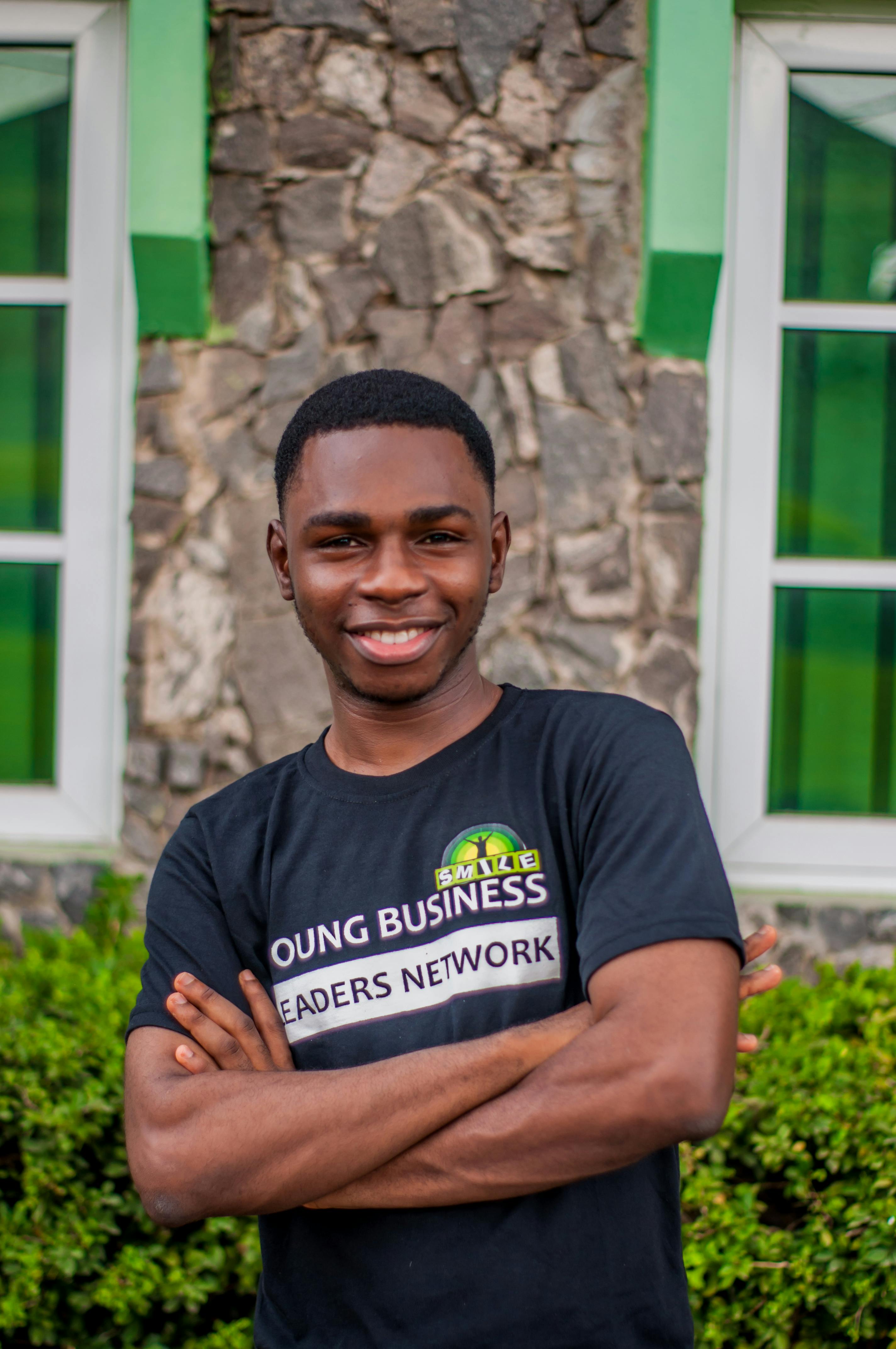 A young man smiles confidently, posing with crossed arms outside a building.