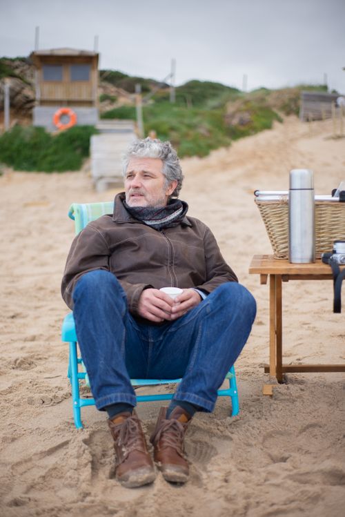 Free Elderly man sitting comfortably on a beach in Portugal, holding a drink, enjoying the outdoors. Stock Photo