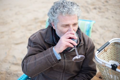 Free Senior man savoring a glass of red wine on a beach picnic. Relaxed and enjoyable outdoor moment. Stock Photo