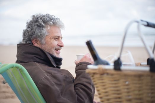 Senior man sitting on a beach with a cup, smiling and relaxing outdoors.