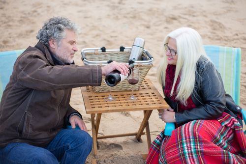 Free A senior couple enjoys a cozy beach picnic with wine on a wooden table. Stock Photo