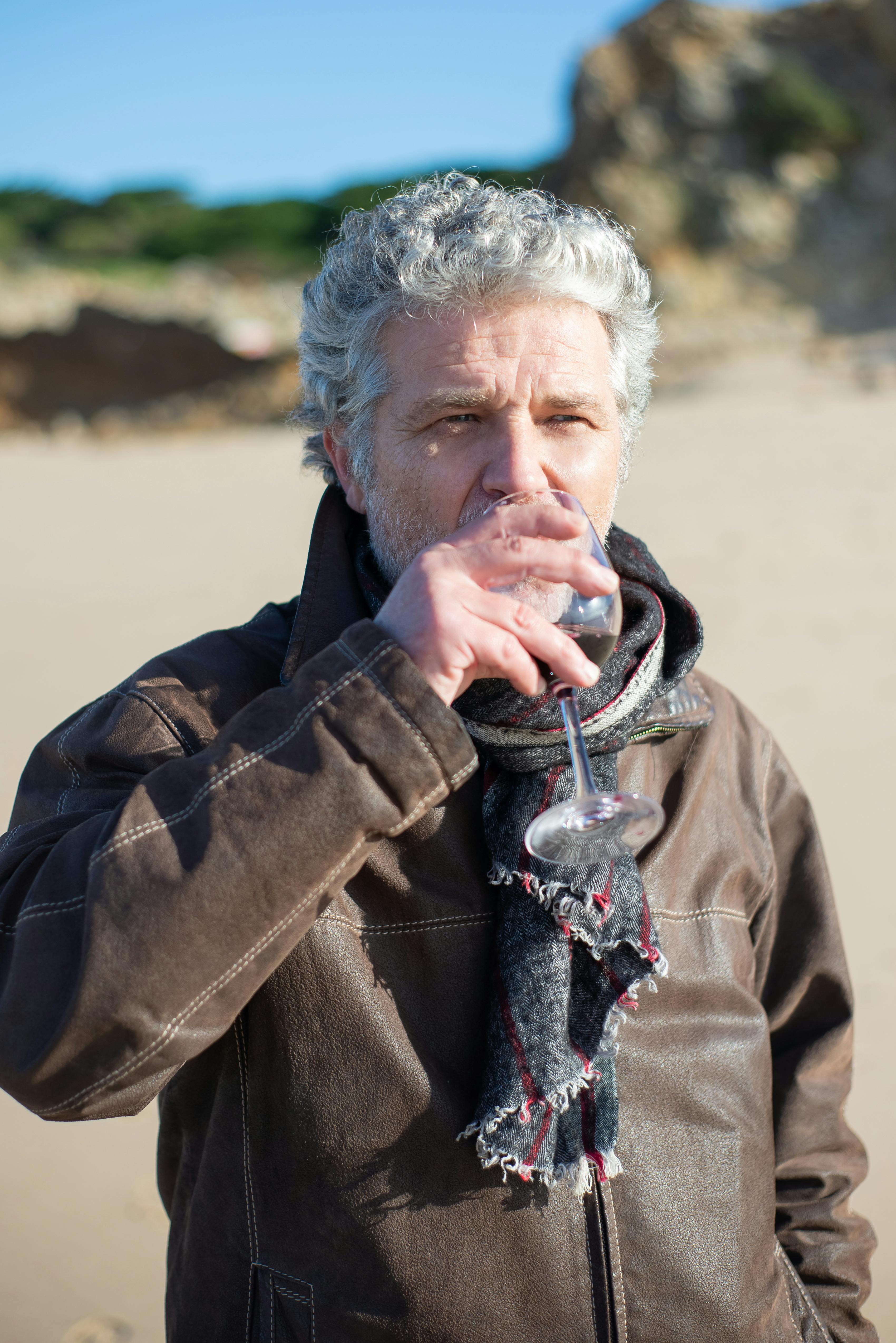 Elderly man in a scarf sipping wine on a sunny day at a Portuguese beach.