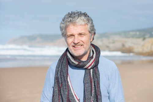 Free A senior man with gray hair and a scarf smiling on a sunny beach in Portugal. Stock Photo