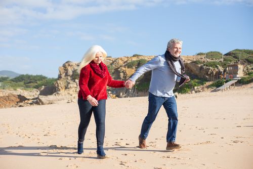 Free An elderly couple holding hands and walking on a sunny Portuguese beach, showcasing love and companionship. Stock Photo