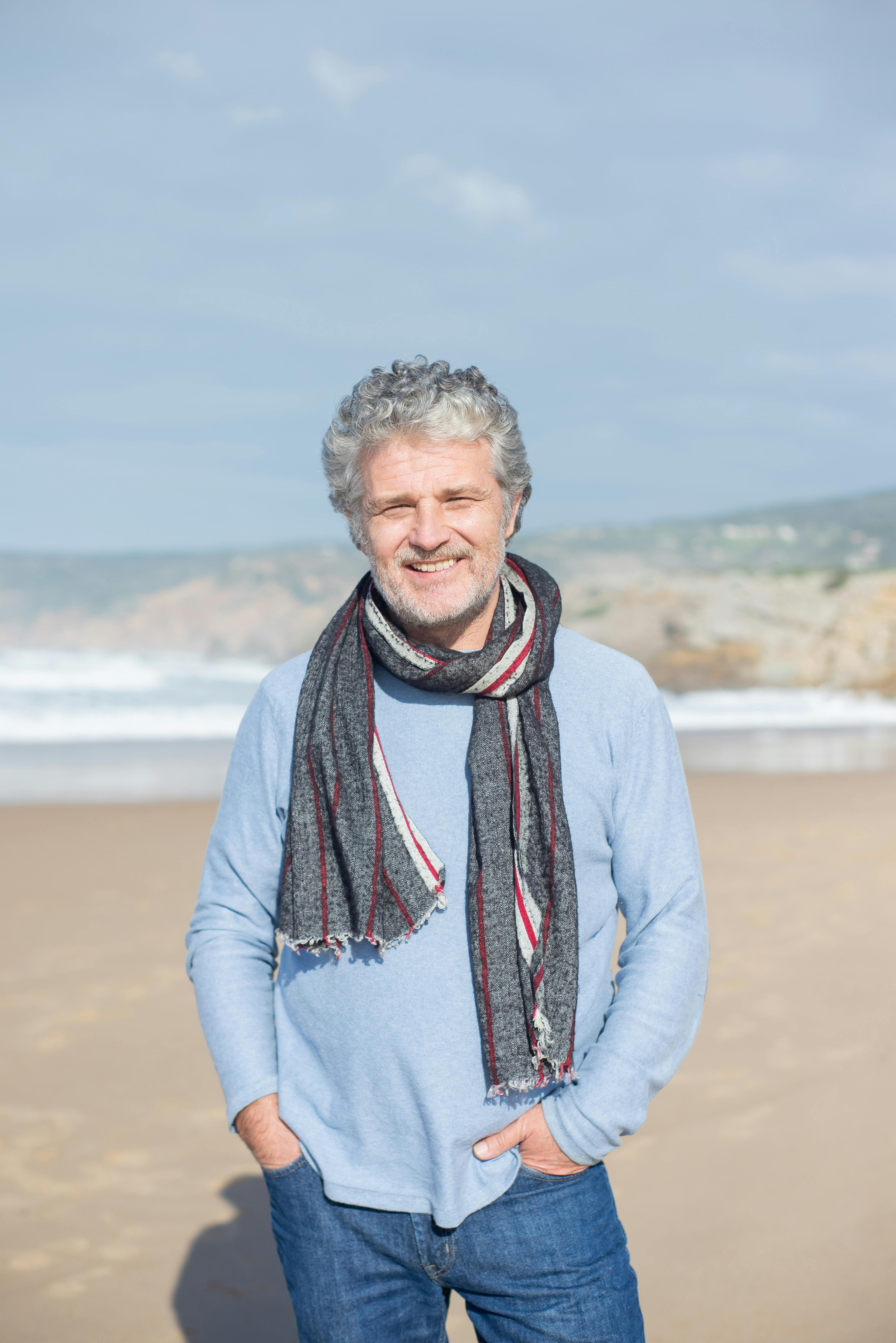 Happy elderly man with gray hair standing on a sunny beach, wearing a scarf and sweater.