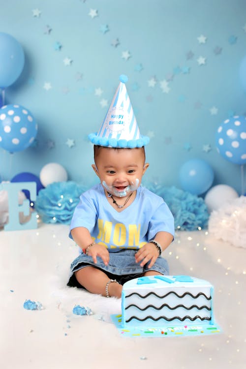 Adorable happy little boy admiring colorful birthday cake among blue balloons in decorated studio