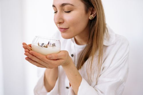 Free A woman in a white shirt smells a floral-scented candle in clear glass. Stock Photo