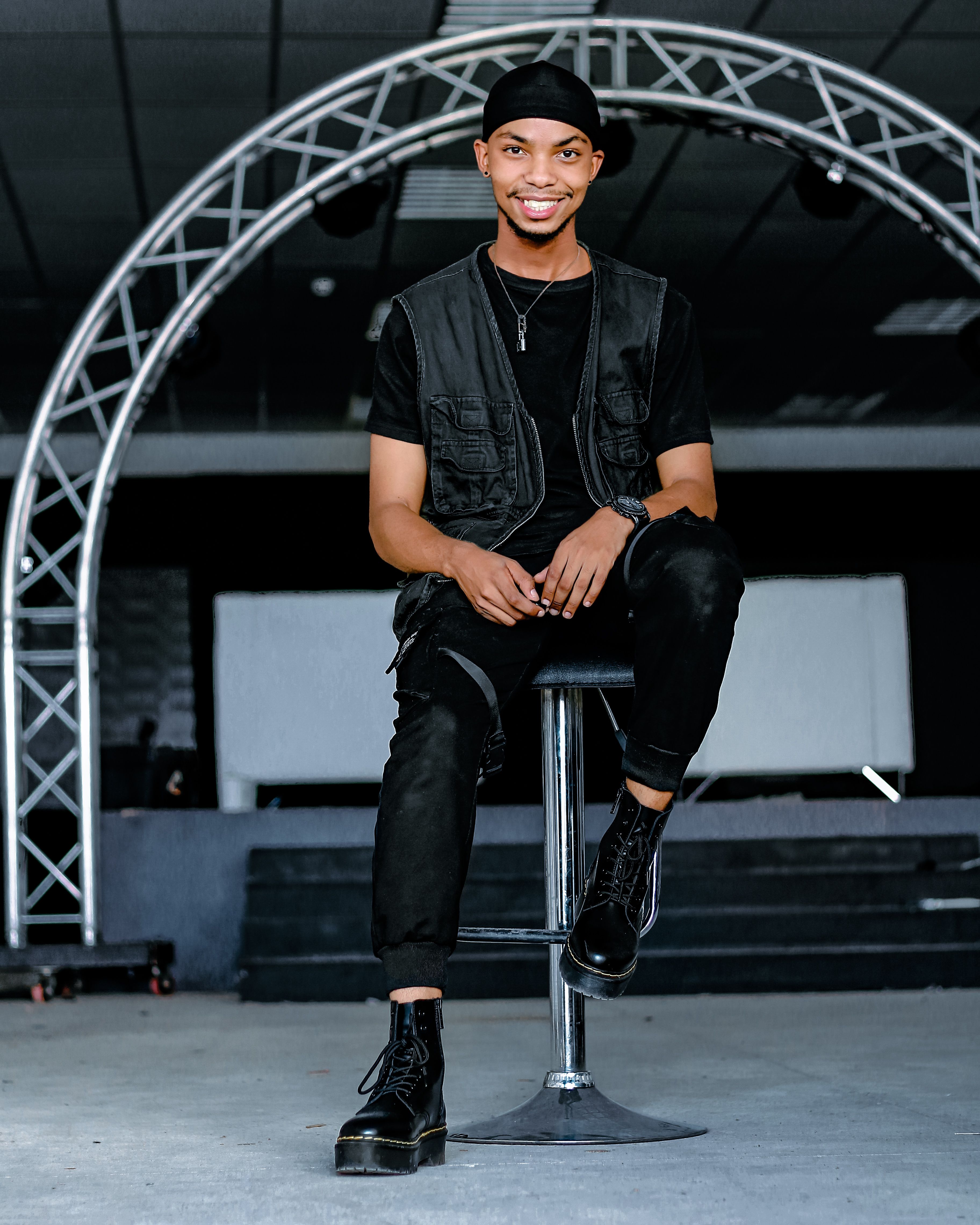 Fashionable young man in black attire sitting on a chair indoors with a big smile.