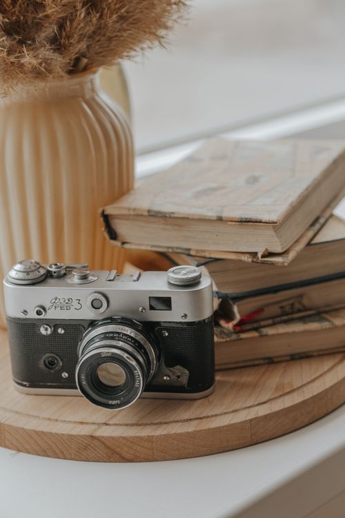 Free Old fashioned photo camera placed on wooden stand near stack of shabby books and vase on windowsill in light room Stock Photo